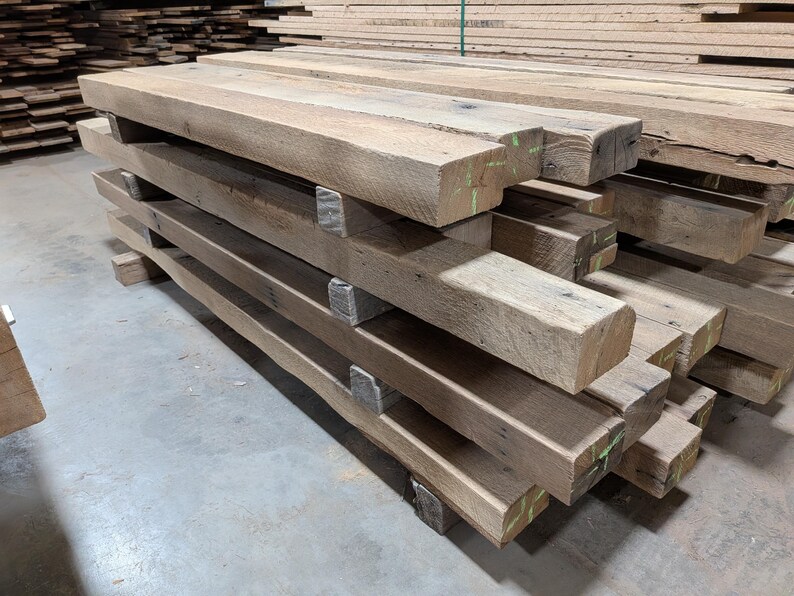 May include: A stack of rough-hewn, rectangular wooden beams. The beams are a natural, light brown color with visible wood grain and some green markings. They are stacked horizontally, supported by smaller wooden blocks.