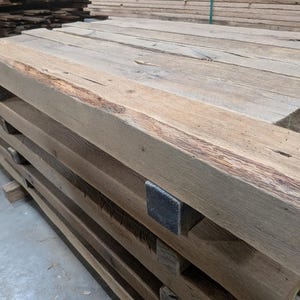 K&ouml;nnte beinhalten: Ein Stapel verwitterter, rechteckiger Holzbalken. Die Balken sind hellbraun mit sichtbarer Holzmaserung und Knoten. Einige Balken weisen dunklere braune Streifen und eine raue, strukturierte Oberfl&auml;che auf. Die Enden der Balken sind rechtwinklig geschnitten.