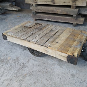 May include: A wooden pallet cart with a weathered, natural wood finish. The cart has a rectangular top with a flat surface and is supported by a metal frame. The cart has four wheels, two of which are visible in the image. The cart is sitting on a concrete surface.