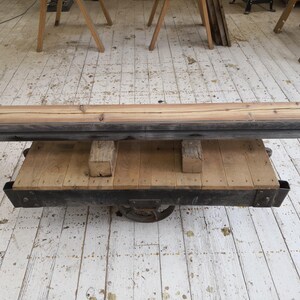 K&ouml;nnte beinhalten: Rustikaler Couchtisch aus Holz mit rechteckiger Oberseite und unterem Regal. Der Tisch hat einen dunklen Metallrahmen und wird von Holzbl&ouml;cken getragen. Die Oberseite besteht aus hellbraunen Brettern, und das Gesamtdesign ist industriell.