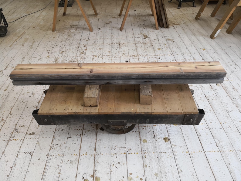 K&ouml;nnte beinhalten: Rustikaler Couchtisch aus Holz mit rechteckiger Oberseite und unterem Regal. Der Tisch hat einen dunklen Metallrahmen und wird von Holzbl&ouml;cken getragen. Die Oberseite besteht aus hellbraunen Brettern, und das Gesamtdesign ist industriell.
