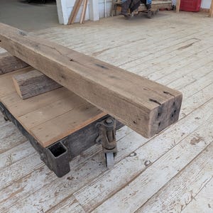 May include: A large, weathered wooden beam rests on a wooden cart with metal wheels. The beam is rectangular, with visible knots and imperfections, suggesting reclaimed wood. Two smaller wood blocks are also on the cart. The cart is on a wooden floor.
