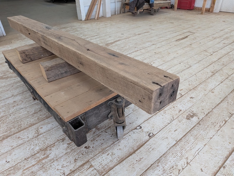 May include: A large, weathered wooden beam rests on a wooden cart with metal wheels. The beam is rectangular, with visible knots and imperfections, suggesting reclaimed wood. Two smaller wood blocks are also on the cart. The cart is on a wooden floor.