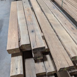 May include: A stack of weathered, rectangular wooden planks with a natural, light brown color. The planks have visible grain and knots, suggesting they are reclaimed or rustic. Some planks have green markings on the ends. The wood appears to be untreated.