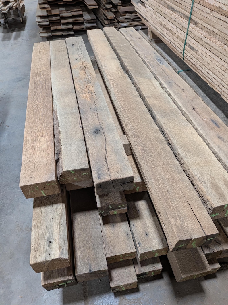 May include: A stack of weathered, rectangular wooden planks with a natural, light brown color. The planks have visible grain and knots, suggesting they are reclaimed or rustic. Some planks have green markings on the ends. The wood appears to be untreated.
