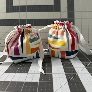 May include: Two small fabric drawstring pouches featuring a white base with a geometric pattern of yellow, pink, dark blue, and teal shapes. Each pouch has a white ribbon closure and a small white tag with the text KNITSPINQUILT on the side.