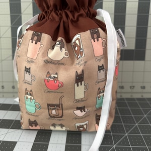 May include: A drawstring bag featuring a repeating pattern of black cats in coffee cups. The bag has a brown top and a tan body with coffee-related text. White ribbon cinches the top. The text "KNITSPINQUILT" is on a white tag.