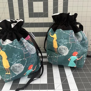 May include: Two drawstring bags with black fabric tops and green fabric bottoms. The bags have a print of women on a chalkboard with equations and a moon.
