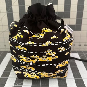 Puede incluir: Bolsa de cordón negro con estampado de taxi amarillo. La bolsa tiene un forro blanco y un cordón negro.