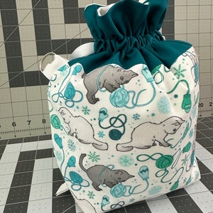 May include: A drawstring bag with a white background featuring a pattern of gray bears, teal yarn balls, mittens, and snowflakes. The top and drawstring are teal, and a white ribbon and metal ring are attached.