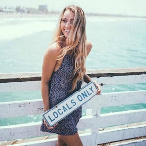 May include: A woman with long blonde hair is standing on a pier holding a sign that says "Locals Only". She is wearing a floral print dress and smiling.