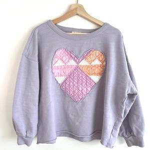 May include: A lavender-colored sweatshirt with a large, patchwork heart design on the front. The heart is composed of various pink, white, and orange fabric pieces, stitched together. The sweatshirt has long sleeves and a crew neck.