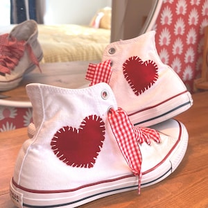 May include: White high-top sneakers with red heart appliqués and red and white gingham laces. The sneakers have a red stripe on the sole and are displayed on a wooden surface.