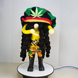 May include: A yellow and green gnome doll wearing a black, red, and green knit hat with a green marijuana leaf design. The gnome has long black dreadlocks and is holding a white cigarette. The gnome is standing on a black rotating display stand.