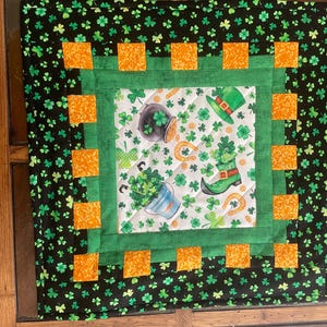 May include: A square quilted fabric featuring a St. Patrick's Day theme. The design includes a central panel with a pot of gold, shamrocks, a green hat, and a green boot. The border is black with green shamrocks and orange squares.