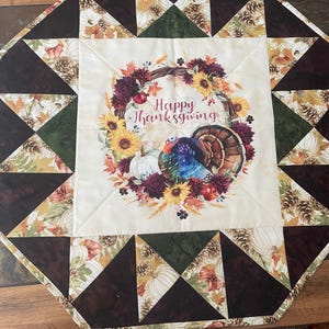 May include: A beige table runner with a patchwork design of brown, green, and white triangles. The center features a wreath with fall foliage, a turkey, and the text "Happy Thanksgiving".