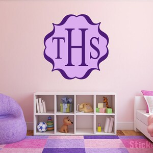 May include: Purple and white monogram decal with the letters "THS" in a decorative frame. The decal is on a pink wall above a white bookshelf with toys and books.