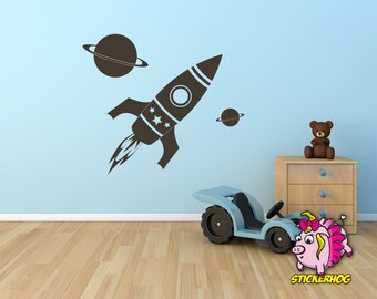 May include: A black and white rocket ship with stars and a ringed planet on a light blue wall. A brown teddy bear sits on a wooden dresser with three drawers. A blue toy car with a pink pig sticker on the floor.