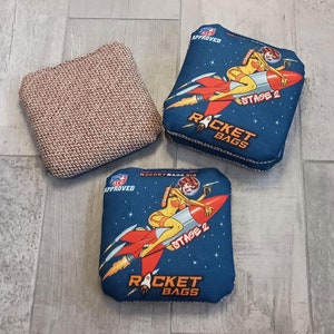 May include: Three square bean bags. Two blue bags feature a retro rocket ship design with a woman in a spacesuit and the text "STAGE 2 ROCKET BAGS". The third bag is a woven brown and tan colour.