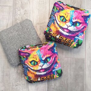 May include: Three cornhole bags. Two bags feature a colorful Cheshire Cat design with the text "Bring It!" and "Rocket Bags." The third bag is a solid gray color. The bags are labeled "ACO Approved."
