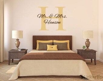 May include: A bedroom with a brown bed with a beige comforter and two pillows. The bed has a headboard with a large gold letter "H" on the wall behind it. The text "Mr. & Mrs. Hanson" is written in black below the letter. There are two nightstands with lamps on either side of the bed.
