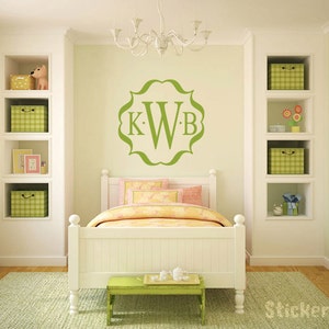 May include: A green monogram decal with the letters KWB in a decorative frame, on a white wall above a white bed with a yellow and pink bedspread in a child's bedroom.