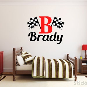 May include: A red letter "B" with black checkered flags on either side and the name "Brady" written in a black cursive font. The decal is on a white wall in a child's bedroom.
