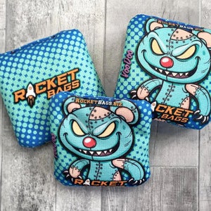 May include: Three blue bean bags with a teal and white polka dot pattern. Each bean bag features a stitched-up monster with a toothy grin and the text "Rocket Bags".