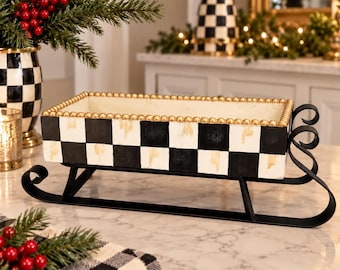 May include: A rectangular sleigh-shaped container featuring a black and white checkered pattern with distressed gold accents. The rim is decorated with a row of small gold beads, and the base is supported by black metal runners with curled ends.