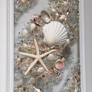 May include: A white framed seashell art piece with a white seashell, starfish, and various other seashells and glass pieces in a clear resin.