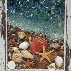 May include: A framed seashell art piece with a blue and white glass mosaic background. The piece features a variety of seashells, including sand dollars, starfish, and scallop shells. The seashells are arranged on a bed of small brown and white stones.