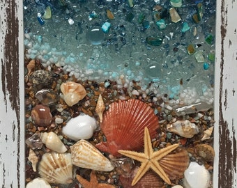 May include: A framed seashell art piece with a blue and white glass mosaic background. The piece features a variety of seashells, including sand dollars, starfish, and scallop shells. The seashells are arranged on a bed of small brown and white stones.