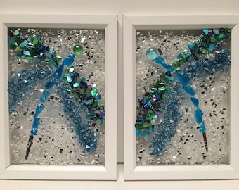 May include: Two framed pieces of art featuring a dragonfly design made with blue, green, and iridescent glass pieces on a white background.