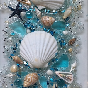 May include: A close-up of a seashell art piece with white seashells, blue glass pieces, and starfish on a white background.