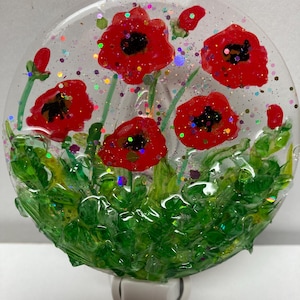 May include: A round glass night light with a red poppy flower design. The night light has a white base and is decorated with green leaves and red poppies. The poppies have black centers and are surrounded by glitter.