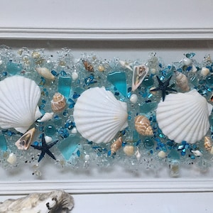 May include: A framed seashell art piece with three white seashells, blue glass pieces, and a starfish on a white background.