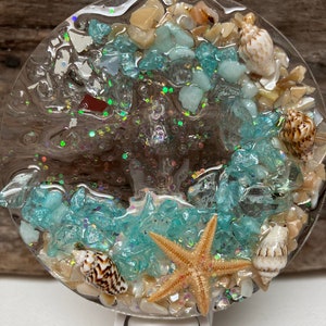 May include: A round night light with a beach theme. The light features a clear resin design with blue and tan stones, seashells, a starfish, and glitter. The base of the light is white.