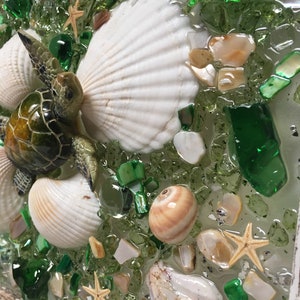 Puede incluir: Una pieza de arte de resina con una tortuga marina, conchas marinas y estrellas de mar en un lecho de piezas de vidrio verde y blanco.