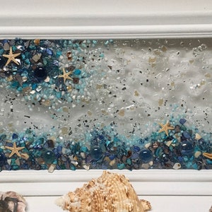 May include: A framed artwork featuring a blue and white seascape with embedded seashells and starfish. The artwork is set against a white background.