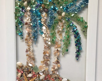 May include: A framed mosaic artwork depicting three palm trees made of seashells and glass beads. The trees are set against a white background and surrounded by a variety of seashells and sand.