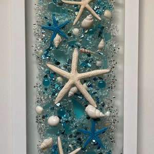 May include: A framed beach-themed artwork featuring a white frame. The artwork showcases a mix of white, blue, and clear elements, including seashells, starfish, and crushed glass. The starfish are in white and blue, and the seashells are in white.