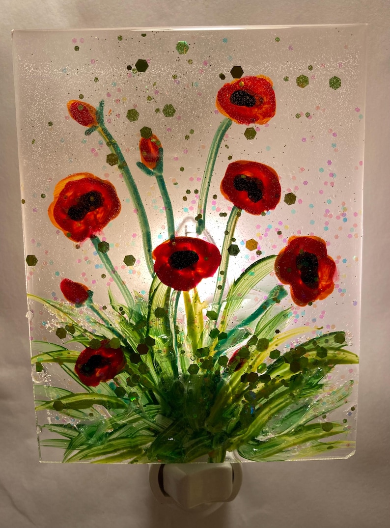 Peut inclure: Une veilleuse avec un motif en verre color&eacute; de coquelicots rouges avec des tiges et des feuilles vertes sur un fond blanc. La veilleuse est branch&eacute;e sur une prise de courant.
