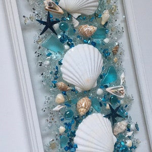 May include: A framed piece of art featuring white seashells and blue glass pieces arranged in a vertical pattern. The background is a light blue colour.