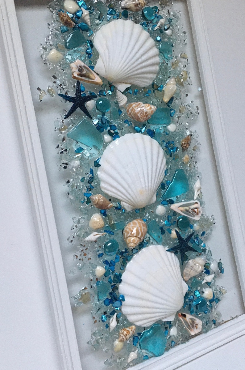 May include: A framed piece of art featuring white seashells and blue glass pieces arranged in a vertical pattern. The background is a light blue colour.