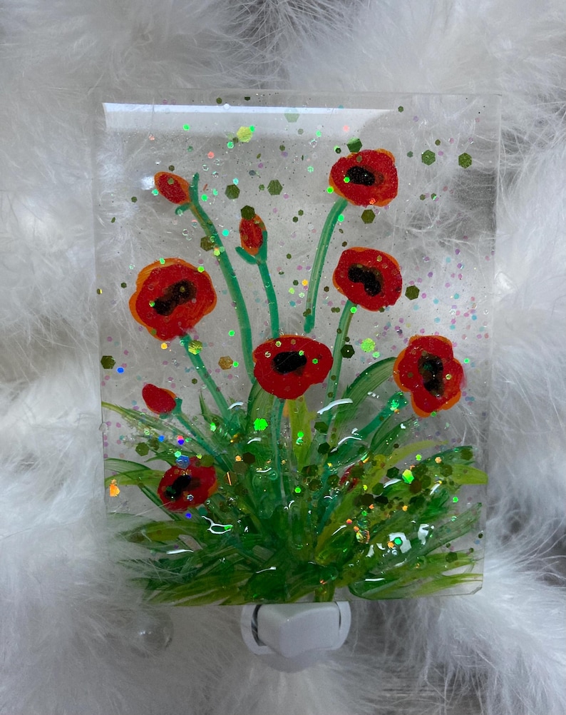Peut inclure: Une veilleuse en verre avec un motif de fleurs de coquelicot rouges. Les fleurs sont peintes sur un panneau de verre transparent avec des tiges et des feuilles vertes. La veilleuse a une base blanche &agrave; brancher.