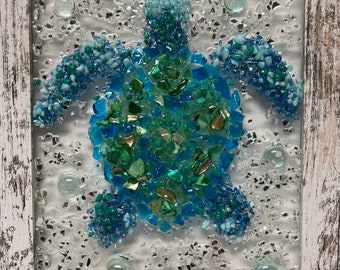 May include: A framed mosaic art piece depicting a sea turtle made of blue and green glass pieces. The turtle is surrounded by small white glass pieces and blue glass beads, creating the illusion of water. A small metal crab is visible at the bottom of the frame.
