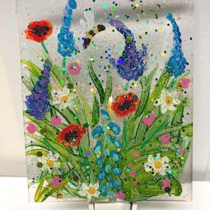 May include: A nightlight featuring a hand-painted scene of a field of flowers with blue, purple, and red flowers, green grass, and a bee. The nightlight is made of glass and has a glittery finish.