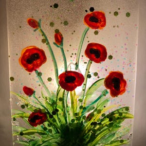 Peut inclure: Un veilleuse en verre teint&eacute; repr&eacute;sentant des coquelicots rouges avec des tiges et des feuilles vertes sur un fond blanc avec des accents de paillettes.