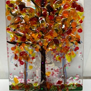 May include: A glass nightlight featuring a colorful autumn tree with red, orange, and yellow leaves. The tree is surrounded by pink glitter and a green base. The nightlight is plugged into a standard electrical outlet.