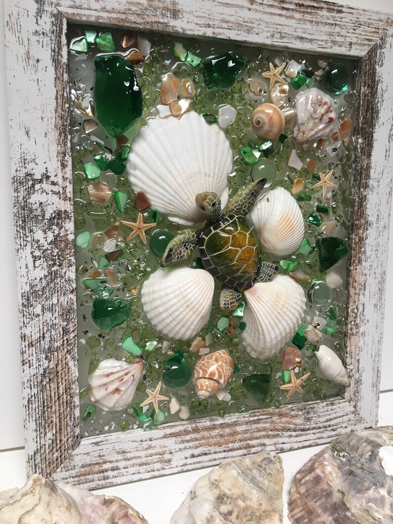 Puede incluir: Una pieza de arte en mosaico enmarcada que presenta una tortuga marina verde rodeada de conchas blancas y piezas de vidrio verde y marr&oacute;n.